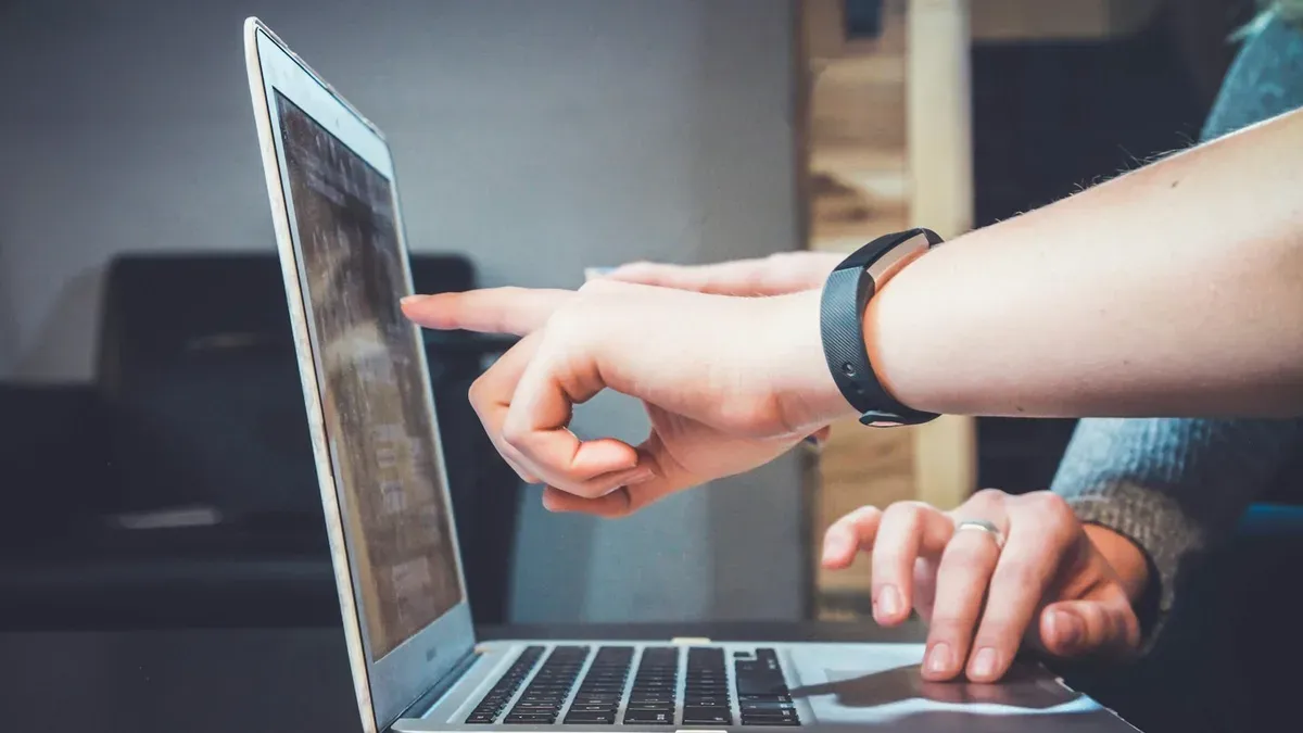 Dwie osoby przy laptopie, jedna wskazuje palcem coś na ekranie, druga trzyma dłoń na gładziku