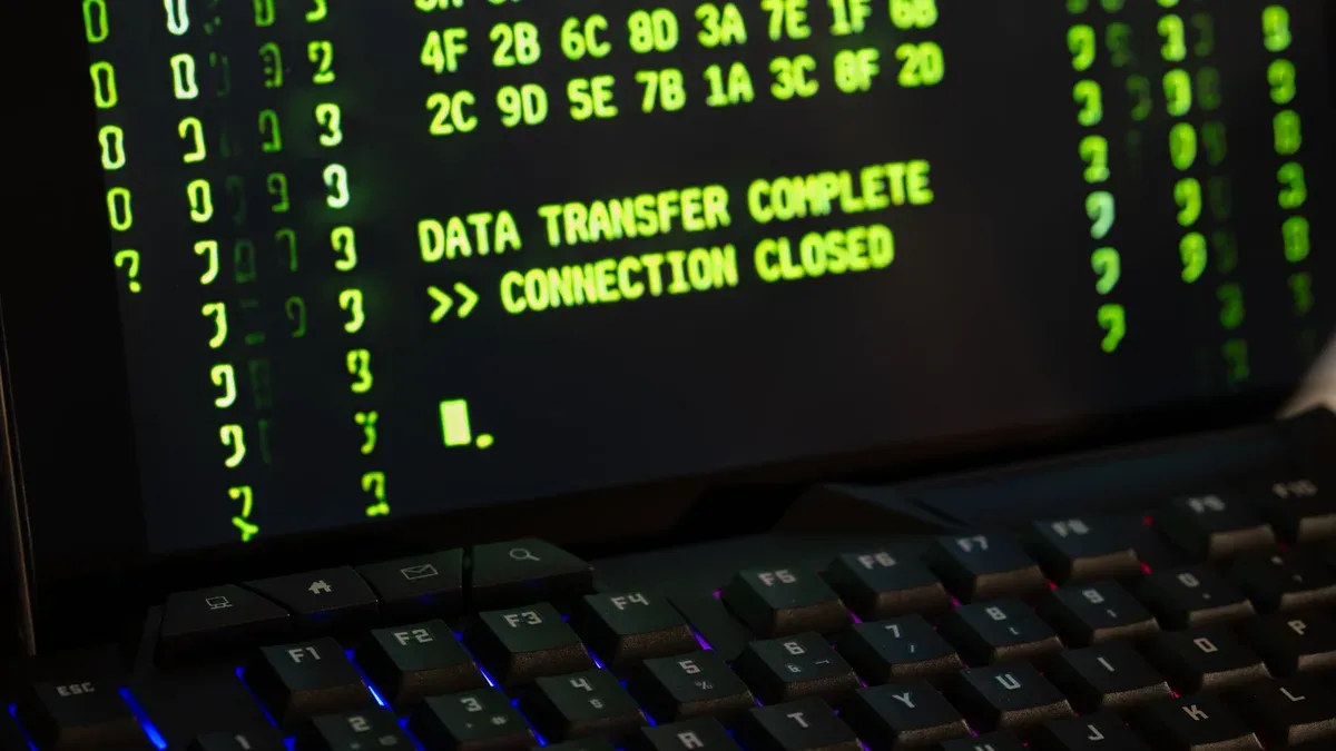 Ekran laptopa z zielonym tekstem w stylu terminala: rzędy kodów szesnastkowych i komunikaty „DATA TRANSFER COMPLETE” oraz „CONNECTION CLOSED”, pod spodem podświetlona klawiatura