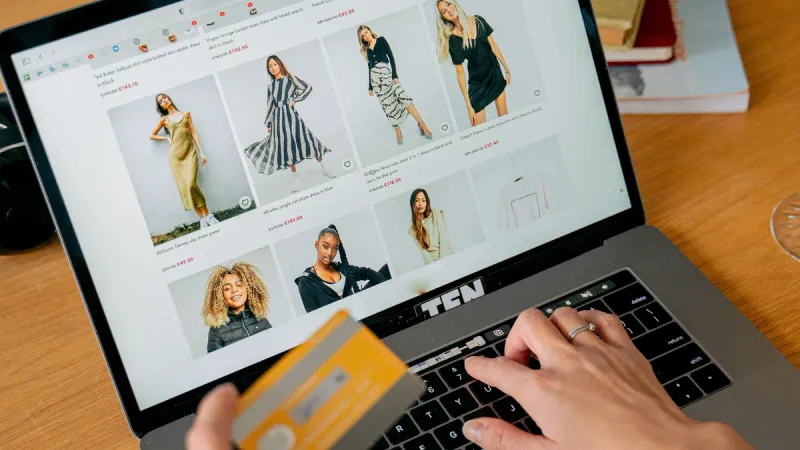 Laptop z otwartą listą sukienek w sklepie internetowym, jedna dłoń na klawiaturze, w drugiej karta płatnicza