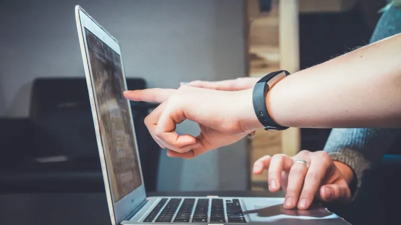 Dwie osoby przy laptopie, jedna wskazuje palcem coś na ekranie, druga trzyma dłoń na gładziku